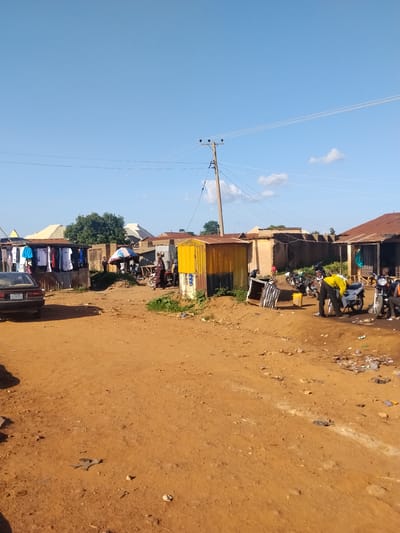 The image depicts a street scene in Mangu Town, Nigeria, characterized by a dusty, reddish-brown dirt road. The environment is informal, with several makeshift structures and stalls lining the road. On the left side, a rack displays various articles of clothing, including t-shirts, being offered for sale. A dark-colored sedan is parked in front of the clothing stall.
Further into the scene, a yellow container-like structure stands prominently, with a corrugated metal roof visible on buildings in the background. A wooden pole with electrical wires stretches across the sky, indicating the presence of electricity infrastructure. Several motorcycles are parked or in use within the scene, suggesting their importance as a mode of transport.
People are present, engaged in various activities. One individual, wearing a bright yellow shirt, appears to be interacting with a motorcycle, possibly repairing it. Another person, dressed in dark clothing, is walking away from the viewer. A child in a red shirt is visible near a building. An umbrella provides shade over a small gathering of people and motorcycles. The overall impression is of a bustling, informal commercial area within a community.