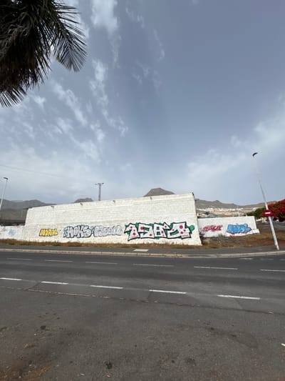 This image depicts a street scene in Adeje, Spain, featuring a white brick wall adorned with graffiti. In the foreground, a paved road with white and yellow lane markings is visible. A portion of a palm tree is in the upper left corner, and the sky is overcast with scattered clouds. The wall itself has various graffiti tags, with prominent ones in yellow, silver, green, and blue. Beyond the wall, a landscape with hills and a few buildings can be seen, suggesting an urban or semi-urban environment. A traffic light and a street lamp are also present.