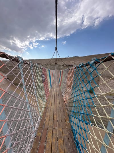 The image shows a suspension bridge with a wooden walkway and brightly colored rope netting on the sides. The bridge extends into the distance, crossing over a dry, rocky terrain. The sky above is partly cloudy with patches of blue sky. In the background, there are some buildings and structures on a hillside. A sign with Chinese characters is attached to the netting on the left side of the bridge. The scene appears to be an outdoor tourist attraction. The specific location is the Jiayuguan Suspension Bridge in the city of Jiayuguan, China.