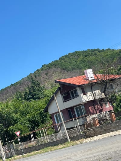 The image depicts a scene in the city of Goritsa, Bulgaria, on a clear day. In the foreground, a paved road is visible, with a dry grassy verge to its left. A low stone and brick wall lines the edge of a property, with a rusted metal fence atop it. A triangular traffic sign with a red border and white interior, indicating a yield or priority road, stands on a pole beside a gate. Beyond the fence, a two-story house with a red tiled roof is situated. The house has maroon and white painted sections and multiple windows. A bare tree with dark branches is in front of the house, and a pink rectangular chimney protrudes from the roof. Behind the house, a densely forested hillside rises, with a section of the forest appearing dry and burnt. The sky is a clear, bright blue.