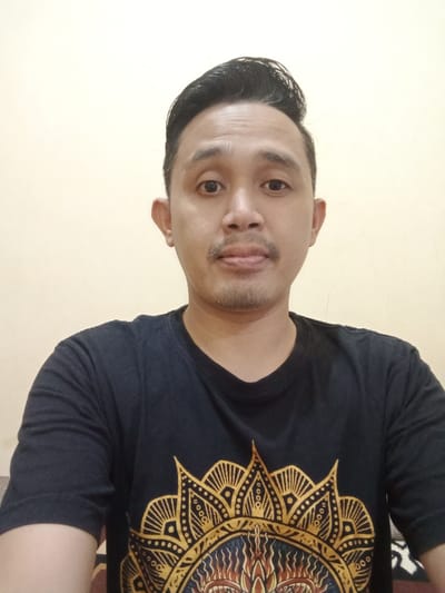 A man with dark hair styled upward, a thin mustache, and a black t-shirt with a golden mandala-like design centered on the chest. He is facing the camera with a neutral expression. The background is a plain, light yellow wall. The man appears to be sitting or standing indoors, with a portion of what looks like a couch visible at the bottom.