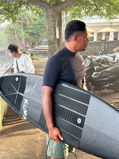 A man in a black wetsuit carries a Kuma surfboard vertically. Another man in a white shirt walks towards the shore in the background. Several motorbikes are parked nearby. The location is Pekutatan, Indonesia.