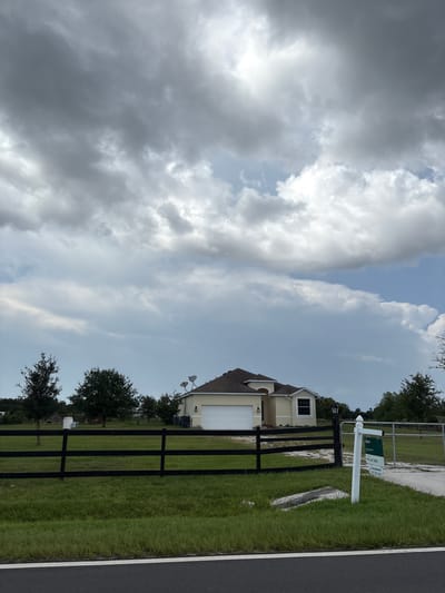 The image shows a yellow house with a white garage door and a black metal fence in front of it. The house is located in Parrish, United States, and there is a "Keyes" real estate sign in front of the fence. The sky is cloudy and there are trees in the background. The house appears to be a single-family residence. The scene is set on a grassy area with a paved road in the foreground. The overall impression is of a suburban or rural residential property.