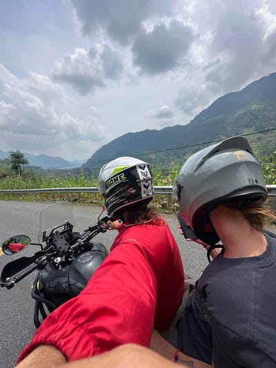 The image displays two individuals stopped with a motorcycle on a mountainous road in Minh Sơn Commune, Vietnam, under a cloudy sky. The original image is rotated, presenting the scene as if viewed from above and to the side, with the horizon running diagonally.
In the foreground, a dark grey motorcycle, possibly an adventure bike with a camouflage-patterned tank, is parked on an asphalt road. One person, wearing a red long-sleeved shirt and a white motorcycle helmet with "YOHE" and black/yellow accents, is positioned near the motorcycle's handlebars. Another individual, dressed in a dark grey t-shirt and a light grey helmet bearing the word "Royal" and an emblem, stands beside them, holding a smartphone. Both individuals appear to be looking downwards or at their phones.
A metal guardrail runs along the edge of the road. Beyond the guardrail, rows of green crops, possibly corn, are visible, cultivated on the slopes. The background is dominated by large, rolling green mountains extending into the distance under an overcast sky filled with prominent white and grey clouds. The scene suggests a break during a motorcycle journey through a scenic, mountainous landscape.