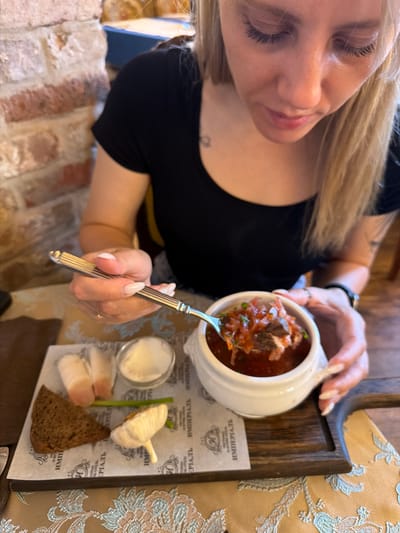 A woman with blonde hair and long fingernails is eating borscht from a white bowl with a fork. She is seated at a table in a restaurant. On the table is a wooden tray with a slice of dark bread, a few pieces of white fatback, a small dish of sour cream, a green onion stalk, and a whole garlic bulb. The background shows a brick wall. This scene takes place in Suzdal, Russia, likely at a restaurant with traditional Russian cuisine.