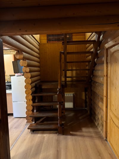 This media file shows an interior view of a rustic wooden home, likely in Novy Gorodok, Russia. The scene features a wooden staircase with carved railings, made from logs and polished wood. The walls and ceiling are also constructed from wood, with visible log beams and planking.

On the lower level, a white refrigerator is visible, along with what appears to be a television screen partially obscured. The overall impression is one of a cozy, traditional dwelling. The lighting suggests it is daytime, with natural light casting shadows from the wooden structures. The style of the home, with its extensive use of logs and natural wood finishes, points to a location that embraces a rustic aesthetic. There is no visible text in the media file.