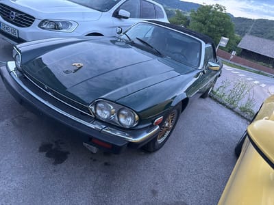 The image displays a dark green Jaguar XJS convertible, equipped with dual circular headlights, chrome trim on the grille and bumper, black rubber bumper accents, and gold-colored wire wheels. A gold leaping Jaguar hood ornament is affixed to the center of the bonnet, which also reflects an overcast sky. A black convertible soft top is in the raised position. Directly beneath the front bumper, an irregular dark fluid stain is present on the asphalt.

To the left and slightly behind the Jaguar, a white Volvo XC60 SUV is partially visible. Its front grille and headlights are prominent, and the Slovenian license plate "CE EI-625" is clearly displayed, indicating registration in the Celje administrative unit, which includes Laško. On the far right foreground, a small portion of a yellow vehicle's bodywork is visible.

The vehicles are parked on an asphalt surface. To the right, white zebra crossing markings are visible on a road. The background features a verdant landscape of tree-covered hills or mountains beneath a cloudy sky, characteristic of the region. A rustic wooden building with a dark, sloped roof and a brown wooden fence is situated in the mid-ground on the right side of the frame. A metal road sign structure is also visible behind the green Jaguar. The ambient light suggests either late afternoon or early evening. The details, including the "CE" license plate prefix, confirm the location as being within the vicinity of Laško, Slovenia.