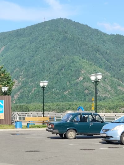 This image depicts a parking lot in the city of Cheremushki, Russia, with a large, forested mountain serving as the backdrop. In the foreground, two cars are parked on an asphalt surface marked with white lines. The car on the left is a dark green sedan, while the car on the right is a light blue hatchback. Behind the cars, there is a wooden bench and blue trash receptacles next to a brown fence. Further in the background, a body of water can be seen, with a white railing along its edge. Multiple lampposts with two lights each are present. The mountain is densely covered with trees, and a telecommunications tower is visible at its peak. The sky is blue with a few scattered clouds. No people are visible in the frame.