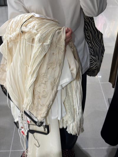 The image depicts a close-up of a person holding several cream-colored garments, likely in a retail setting. The garments are layered and appear to be made of delicate fabrics, with some featuring intricate embroidery and lace details. One garment has a ruched or gathered waistband, and another displays a floral embroidered pattern. Price tags are visible on some of the items. The person is wearing a white shirt, and a bag with a black and white zebra print is partially visible. The background suggests an indoor environment with tiled flooring and hints of other clothing displays. The scene is located in the city of Santiago De Compostela, Spain, possibly within a clothing store near the cathedral.