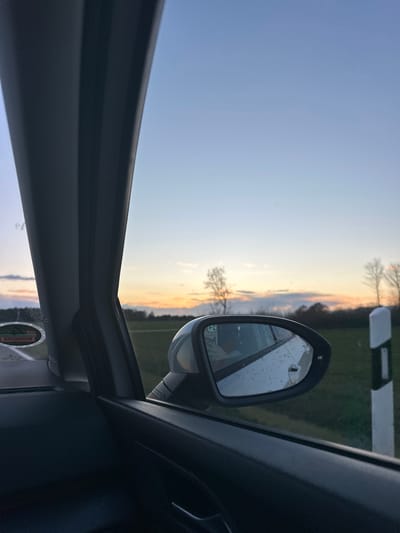 The image shows the view from inside a car, likely on a road near Haundorf, Germany, at sunset. The sky is a gradient of blue to orange. A side mirror is visible, reflecting the sunset and a roadside marker. The car's interior is dark, and a portion of the window frame is in view. The location is inferred from the presence of a roadside marker typical of Germany, and the general landscape is characteristic of the region around Haundorf. There are no people visible.