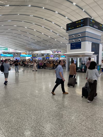 The image depicts the interior of a modern transportation hub, likely an airport or a high-speed train station, in Longhua District, China. The architecture features a sweeping, curved ceiling with integrated lighting, creating a spacious and illuminated environment. In the foreground, several individuals are visible walking across a polished, speckled floor, some with luggage. To the right, a tall, multi-sided signage tower displays directional information in both Chinese characters and Arabic numerals, indicating gates and platforms such as "8B-20B", "6A-7A", and "B6-B7". In the middle ground, a large waiting area is populated with numerous people seated on rows of benches, facing digital display boards that likely show schedules or travel information. Further in the background, more seating areas and information displays are discernible, along with glimpses of the exterior through large windows. The overall scene conveys a sense of organized transit activity.