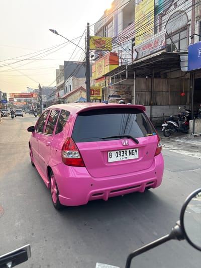 The image depicts a bright pink Honda Jazz car driving down a city street in Kelapa Dua, Indonesia. The car is the primary subject, viewed from the rear-left quarter. It has a spoiler and custom pink rims. The license plate reads "B 1939 NGY". The street is paved with asphalt and lined with buildings on both sides. These buildings house various businesses, indicated by signage such as "FEDERAL OIL CENTER" and "DINO DINO", suggesting a commercial area. Power lines are strung overhead, and street lamps are visible. Other vehicles, including motorcycles and cars, are present on the road, indicating moderate traffic. The lighting suggests it is late afternoon or early evening, with a soft, warm light casting long shadows. The overall environment appears to be an urban commercial street.