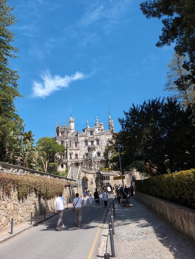 The image shows a section of Quinta da Regaleira in Sintra, Portugal, under a clear blue sky with scattered cirrus clouds. The palace complex, known for its elaborate architecture, is visible to the left. The perspective is from a slightly lower angle, looking upwards towards the building's spires and decorative elements.
A paved pathway runs along the right side of the frame. A group of people is walking along the pathway. The path is bordered by a stone wall topped with shrubbery on the left and a higher stone wall on the right, separating it from a cobblestone walkway.
The vegetation is lush and green, with trees framing the top and bottom of the image. A stone balustrade separates the garden from the pathway.