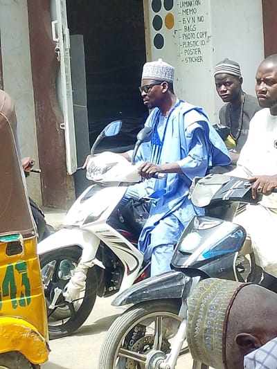 The image captures a street scene in Zango, Nigeria, under daylight conditions. A man is prominently featured, seated on a white JINCHENG motorcycle with black accents. He wears a light blue traditional kaftan, a white patterned cap, and black sunglasses, with his hands positioned on the handlebars. To his right and slightly behind him, another man with a dark complexion, wearing a dark cap and black shirt, observes the scene. A third man, partially visible to the right, wears a white garment and displays a ring on his right ring finger while holding handlebars of an unseen vehicle. In the far left foreground, part of a yellow tricycle marked with "43" in green is visible. Another motorcycle, black with "DONE" visible on its silver rims, is partially present in the lower right. The background consists of a building with an open doorway and a white sign detailing services: "LAMINATION," "MEMO," "BVN NO," "BAGS," "PHOTO COPY," "PLASTIC ID," "POSTER," and "STICKER," with three black and one yellow circular elements above the text.