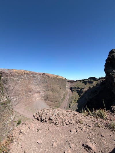 This is a wide-angle photograph of the crater of Mount Vesuvius, located near Ercolano, Italy. The image is dominated by the vast, barren expanse of the volcanic crater under a clear, bright blue sky. The crater walls are steep and composed of reddish-brown volcanic rock, with visible layers and textures indicating geological formations.

In the foreground, the edge of the crater is visible, covered in loose volcanic soil and scattered rocks. Sparse vegetation, including some dry grasses and small plants, grows on the slopes and near the rim. The interior of the crater shows a conical deposit of ash and debris in the center, with gentler slopes leading up to the higher rim.

The lighting suggests it is daytime, with the sun casting shadows within the crater, highlighting its depth and rugged terrain. The overall impression is one of natural grandeur and geological power. There are no people or man-made structures immediately visible in the crater itself, emphasizing the natural environment.