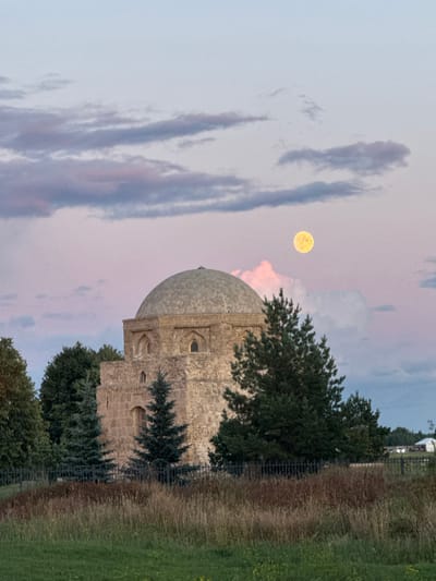 The image depicts a stone building with a domed roof, likely a historical monument, in the city of Bolgar, Russia. The building is constructed from rough-hewn stones and features arched windows. It is surrounded by trees, including evergreen conifers and deciduous trees. A tall, full moon, appearing yellowish, is visible in the pale pink and blue sky, which is streaked with thin clouds. The foreground consists of dry grass and vegetation, separated from the building by a dark, wrought-iron fence. In the background, more trees and a white structure, possibly a building with a roof, are visible, along with what appears to be a car. The overall scene suggests a quiet, possibly rural, setting at dusk or dawn.