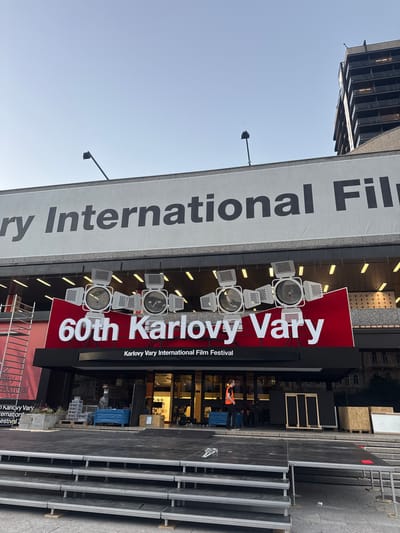 This media file captures an exterior view of a building adorned with a large banner promoting the "60th Karlovy Vary International Film Festival". The banner, primarily white with bold black lettering, displays "ry International Fil" on the left. To the right, a vibrant red section features the text "60th Karlovy Vary" in large white font, with "Karlovy Vary International Film Festival" written below in a smaller font.

Several large, studio-style spotlights are mounted on the building facade, positioned above the "60th Karlovy Vary" text, suggesting an outdoor setup for an event. In the background, through glass windows, some interior lights are visible, and a person wearing an orange vest can be seen. The scene also shows stacked black platforms or stages to the right of the building, indicating preparations for an event or festival.

The sky above is clear and a pale blue, suggesting it is daytime, likely morning or late afternoon given the angle of the light. The overall impression is one of anticipation and preparation for a significant cultural event. The context provided identifies the location as Karlovy Vary, Czechia.

Visible text includes "ry International Fil", "60th Karlovy Vary", "Karlovy Vary International Film Festival", and partial text "ary", "-estival", "Kariovy Vary", "ernat on", "estival 2" on a lower banner or sign.