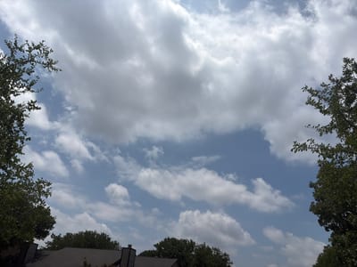 A medium shot captures a partly cloudy sky over a residential area in Grapevine, Texas. Two mature trees, one on the left and one on the right, frame the sky, their branches reaching towards the center. Below the trees, the dark, sloped roofs of houses are visible, punctuated by chimneys. The clouds are a mix of fluffy white cumulus and larger, gray formations, indicating varied atmospheric conditions. The sky is a light blue, with sunlight peeking through the clouds. The scene is a typical suburban landscape on a day with dynamic weather.