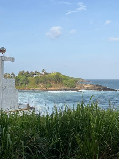 The image shows a view of the ocean and a rocky coastline with lush vegetation in Talalla, Sri Lanka. In the foreground, tall green grass dominates the lower portion of the frame. To the left, a white structure with a cross shape on top is partially visible, with a satellite dish mounted on its surface. The middle ground features a body of water with visible waves breaking against the shore. Beyond the water, a green, hilly landscape is covered with palm trees and other foliage, with a few buildings peeking through the greenery. The ocean extends to the horizon, meeting a clear blue sky with a few wispy clouds. The overall scene depicts a tropical coastal environment.
