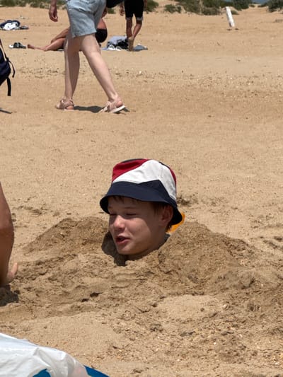 The image depicts a sandy beach in Vityazevo, Russia, with several individuals present. In the foreground, a young boy wearing a red, white, and blue bucket hat is partially buried in the sand, with only his head and shoulders visible. He appears to be looking towards his left with his mouth slightly open. To the left of the boy, the leg of another person, wearing light-colored shorts and pink sandals, is visible walking away from the camera. In the background, further up the beach, other beachgoers can be seen, along with some scattered clothing and beach accessories. The environment is characterized by sand and sparse vegetation in the distance, suggesting a sunny day at the beach.