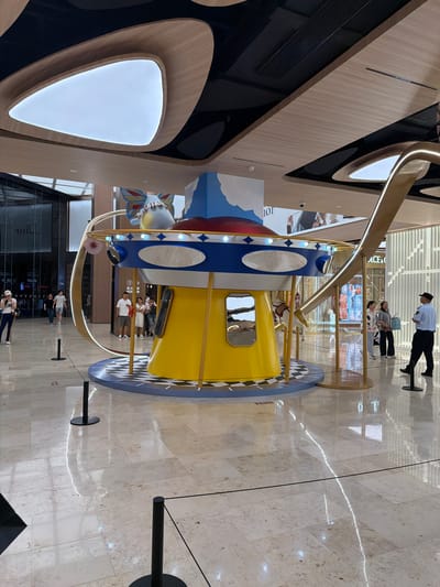 The image captures a brightly lit indoor scene, likely a shopping mall in Xuanwu District, China, given the context. The dominant object is a colorful, carousel-like structure with a yellow and blue cylindrical cabin, adorned with red and white circular patterns. It appears to be a children's ride or a decorative installation, featuring a whimsical design with a large, cartoonish character on one side and a large golden, curved beam above it.
Several people are visible in the frame. In the background, a security guard in a white shirt and black pants stands near the carousel, observing. A group of individuals, likely shoppers, are also present, some standing near the ride and others walking by. Closer to the foreground, another group of people are seen walking on the polished marble floor. One person is holding a phone and appears to be taking a picture.
The setting is spacious and modern, with sleek wooden paneling on the walls and a reflective marble floor that gleams under the bright overhead lighting. The overall atmosphere seems to be one of leisure and commercial activity, typical of a shopping mall. The time of day is not explicitly indicated but the bright indoor lighting suggests it's during operating hours. There are no obvious indicators of the weather.
Visible text includes "DIOR" on a storefront in the background, suggesting a high-end retail environment. The context provided about Xuanwu District, China, further localizes the scene. The mood is neutral, depicting a typical day in a public space with shoppers and a central attraction.