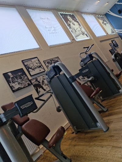 The image depicts the interior of a gym in Oberseelbach, Germany, devoid of occupants. The scene is dominated by multiple Technogym brand fitness machines. In the foreground, a grey and black Technogym bicep curl or similar arm isolation machine with brown upholstered seating and chest pads is visible. Behind it, another grey and black Technogym strength training machine, featuring a cable system and brown upholstered pads, is partially visible. Further to the right, a third, largely obscured Technogym machine stands.

The walls are light-colored. The upper section features four rectangular, illuminated panels. The two leftmost panels are uniformly white with faint handwritten text on the second. The third panel from the left displays a black and white image of an athlete, while the fourth on the far right is partially visible. Below these, a cluster of at least five framed black and white photographs depicting sports scenes, including what appears to be ice hockey and basketball, are mounted on the wall.

The floor is covered with light brown, wood-effect laminate. The ceiling consists of white acoustic tiles, with a blue and white wave-like design element visible on the upper right side. The overall lighting is consistent with an indoor environment.