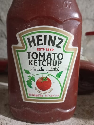 A close-up shot captures a Heinz Tomato Ketchup bottle against a neutral, light-colored background that appears to be a wall or countertop. The bottle is predominantly red with a prominent label featuring the Heinz logo, the words "TOMATO KETCHUP," and some text in Arabic. A tomato with leaves is depicted on the label, and the words "GROWN NOT MADE" and "Net Weight 1kg" are visible. The bottle itself shows some dust or residue on its surface. The overall scene is static and focuses entirely on the product. The provided context about the city of Taraclia, Moldova, does not correspond to any visual information within the image itself.