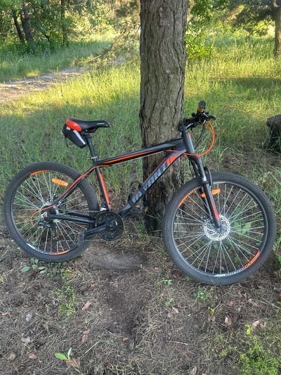 A black Avanti brand mountain bike with orange accents is leaning against a tree on a dirt path in a wooded area. The bike has disc brakes on both wheels, a black seat with orange accents, and a small black bag attached to the seat post. The surrounding environment is characterized by green grass, trees, and pine needles on the ground, suggesting a natural, possibly rural setting. The lighting indicates it is daytime, with sunlight filtering through the trees. The scene appears to be located in the city of Chmyrivka, Ukraine.