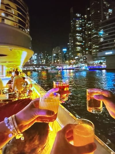 In this media file, a group of people are enjoying drinks at night, likely on a boat or a waterfront venue, given the reflections of city lights on the water. The scene is illuminated by artificial lights from the city skyline and some internal lighting.
The primary subjects are hands holding clear glasses filled with amber and yellowish beverages. One hand wears an ornate bracelet with charms, suggesting a celebratory or social occasion. In the background, out of focus, are glimpses of other people and what appears to be a boat's railing or seating area. The reflections in the water suggest a metropolitan setting with tall buildings.
The activity is a toast or a casual gathering where drinks are being shared. The time of day is clearly night, with the city lights creating a vibrant backdrop. The mood appears relaxed and social. The location context is Pori, Finland, which might imply a summer evening given Pori's known festivals and long daylight hours during summer. However, the image itself predominantly shows nighttime. There is no visible text in the media file.