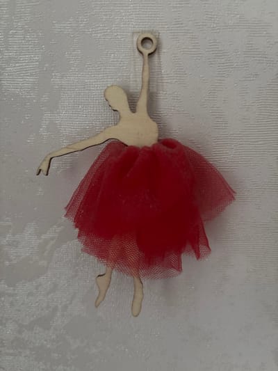 This is a close-up, eye-level, indoor, still photograph of a wooden ballerina ornament with a red tulle tutu. The ornament is positioned slightly left of the center of the frame, against a white, textured surface that appears to be fabric or wallpaper. The ballerina's figure is light brown wood and depicts a dancer in a classic ballet pose with one arm extended and the other bent. She is wearing a vibrant red tutu made of layered tulle fabric, which is full and flares out from her waist. The background has a subtle, embossed pattern. The lighting is even, and there are no shadows visible. The scene is static and does not depict any activity or interaction. The location is not discernible from the image itself.