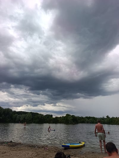 The image depicts a cloudy day at a lake in Neckartenzlingen, Germany. In the foreground, a sandy beach is visible, with a colorful inflatable boat resting on the sand. Several people are in the water. On the right, a man in light green shorts stands at the water's edge, facing away from the camera. Further out in the lake, people are paddleboarding and swimming. The background is lined with lush green trees, forming a natural border around the body of water. The sky is dominated by dramatic, dark storm clouds, suggesting an impending weather change.