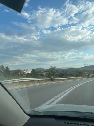 An outdoor, daytime, eye-level shot captures a view from inside a vehicle on a paved road. The road, marked with double white lines, curves slightly to the right. A metal guardrail runs along the left side of the road, separating it from a dry, grassy embankment. Beyond the embankment, the landscape unfolds with rolling hills covered in scattered vegetation and trees. In the middle ground, a modern, white building complex with several units and a red car parked in front is visible, situated amidst greenery. Further in the distance, a large, pylon-like structure with power lines is prominent against the sky, and undulating hills fade into the horizon under a partly cloudy sky. The sky itself is a vibrant blue, interspersed with scattered white and gray clouds. The lower portion of the image shows the interior of the vehicle, including the dashboard and windshield.