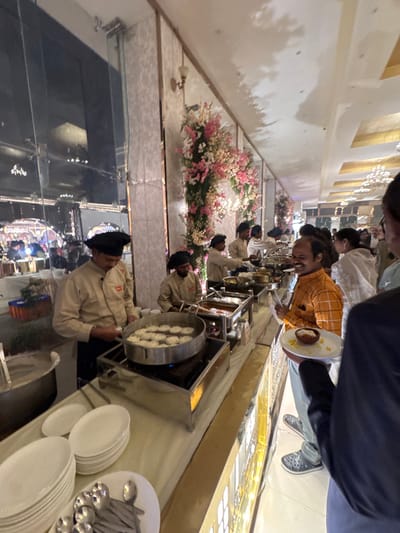A buffet line is set up, likely for a wedding or large event. A chef fries food in a large pan. Guests line up to serve themselves, filling their plates with various dishes. Pink and white flowers decorate the table. Plates and serving spoons are stacked on the edge of the table. The venue appears to be a brightly lit hall with patterned walls.