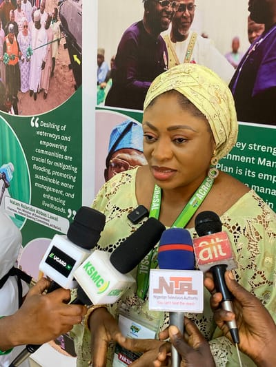 A woman in a light yellow-green lace top and a matching head covering is featured prominently, looking slightly downwards with a focused expression. She is surrounded by several hands holding microphones towards her, indicating a press conference or interview. She wears a green lanyard with "ACRESAL" printed on it.
The microphones display logos of various media outlets: AIT, NTA (Nigerian Television Authority, with the slogan "You can't beat the reach"), KSMC, and UGARD TV. The setting appears to be an indoor event, likely in Kwarau, Nigeria, given the visible Nigerian media outlets.
In the background, a large banner is partially visible, featuring text and images of other people. Notable text on the banner includes a quote attributed to "Malam Balarabe Abbas Lawal, Hon. Minister of Environment, Chairman, Federal Steering Committee, ACRESAL": "Desilting of waterways and empowering communities are crucial for mitigating flooding, promoting sustainable water management, and enhancing livelihoods." Other fragments like "ACRES" and "Agro-Olma" are also present on the banner, further indicating the context of the ACRESAL project. The image is taken from a slightly elevated perspective.