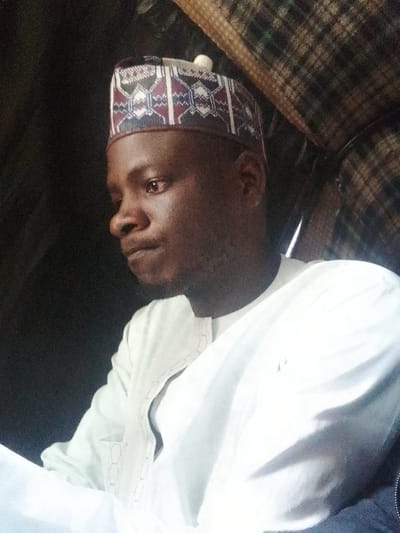 The image shows a close-up, profile view of a young man from Unguwar Saya, Nigeria. He is wearing a traditional cap with geometric patterns and a white garment with intricate embroidery on the collar. The man is looking to his left, with his head slightly tilted. The background is dark, with some textured fabric visible in the upper right corner, suggesting he might be inside a vehicle or a covered structure. The lighting is dim, highlighting the man's face and the details of his attire.