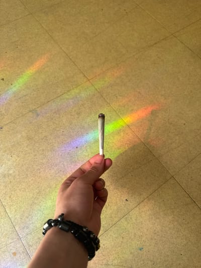 The image shows a person's right hand holding a lit, hand-rolled cigarette, commonly known as a joint. The cigarette has a dark, ash-covered tip, indicating it is burning or recently smoked. A black beaded bracelet is visible on the wrist. The scene takes place indoors, with the hand and cigarette positioned above a light-colored, possibly beige or yellowish, tiled floor featuring distinct grout lines. Prominent, colorful light reflections, resembling a rainbow spectrum (including red, orange, green, blue, and purple hues), are cast across the floor. These reflections suggest sunlight passing through a prism, stained glass, or a similar object, indicating that the photo was taken during daytime, likely on a sunny day. No explicit location cues are visible within the image to confirm the provided context of Orsay, France. There is no visible text or discernible emotional expression.