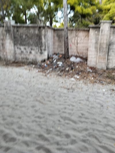 The image depicts an outdoor scene in Gombato Bongwe ward, Kenya. The foreground consists of a light-colored, granular surface, appearing to be sand or loose dirt with visible undulations and textures. Moving towards the background, a distinct line marks the beginning of an area densely covered with assorted refuse, including plastic items, wrappers, and general litter, intermingled with dried leaves and sparse brown vegetation. A vertical, slender tree trunk rises from this littered area, positioned off-center to the right. Behind this, a weathered concrete or masonry wall extends horizontally across the frame, featuring lighter-colored sections and darker, possibly stained or dirty patches, with several integrated vertical pillar-like structures. Above and behind the wall, blurred green foliage from multiple trees is visible against a lighter sky, indicating an vegetated area beyond the wall. No people, specific actions, or direct interactions are discernible within the frame. The overall scene suggests an area impacted by waste accumulation.