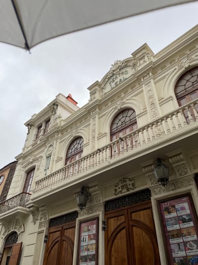 An upward-angled, slightly rotated view captures the ornate facade of the Teatro Leal in La Cuesta, Spain, under an overcast sky. The grand building is cream-colored and features classical architectural elements, including decorative carvings, balustrades, and rounded windows with intricate wooden grilles. Two large, dark wooden doors are visible on the right, flanked by elegant lanterns.
Visible text "TEATRO LEAL" is prominently displayed on the building's facade, confirming its identity. The year "1915" is carved into the stonework, likely indicating its construction date. Advertising posters for upcoming events are affixed near the entrance. These posters display "ABRIL PROGRAMACIÓN 2023" and "ABRIL PROGRAMACIÓN 2026," listing various performances such as "Superthings," "Santi Rodriguez," "Kike Pérez," "Sun Dance Family Sessions," "La Urbana," "Cristina Ramos," "Veladas Infantiles," "Veladas - Ghibli," "Jay Kalo," "II Feel the Talent," and "MASTERCLASS."
A large, white or light-colored umbrella is partially visible in the bottom-left foreground, suggesting the photographer might be sheltered from rain or sun. The scene is quiet, with no people actively engaging in events, but the posters indicate cultural activities take place there. The grey, cloudy sky suggests daytime, possibly a cool or damp day.