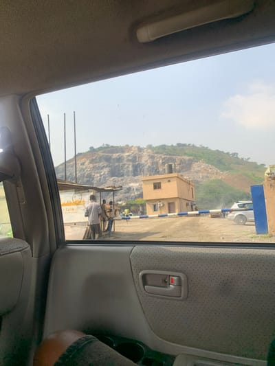 The image is taken from inside a vehicle, looking out at a quarry site in Dam Camp, Nigeria. The sky is clear and bright, suggesting it is daytime. The scene features a large, rocky cliff face, characteristic of a quarry. Below the cliff, there is a small, light-colored building with windows and an air conditioning unit. Nearby, a white vehicle is parked, and several people are visible. One person, wearing a white shirt, is standing in front of a structure with signage. Another group of people appear to be engaged in some activity near a truck. The interior of the vehicle is also visible, showing the car door panel with a handle, and part of the seat. The overall impression is of an active industrial site.