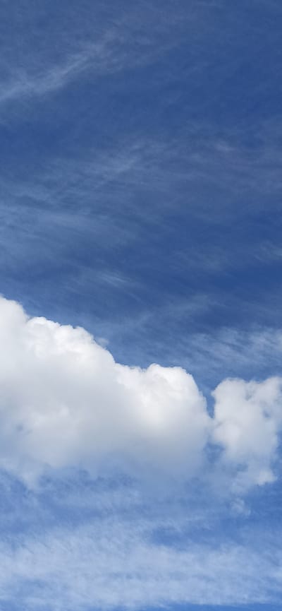 This is a vertical, high-angle photograph of a bright blue sky filled with wispy white clouds. A large, fluffy cumulus cloud dominates the right side of the frame, its edges illuminated by the sun. The left side of the sky features thinner, more streaky cirrus clouds that create a textured appearance. The overall impression is one of a clear and pleasant day, likely late morning or early afternoon given the brightness of the sky. There are no visible people, objects, or text in the image. The scene is a natural sky formation, suggesting an outdoor setting in a location with clear atmospheric conditions. The provided location context of Asbest, Russia, does not offer further visual cues within the image itself.
