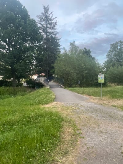 A walking path curves through a park-like area with lush green grass and trees. On the left, a small arched bridge crosses a waterway, with buildings visible beyond the trees. On the right, a signpost with a green sign is planted in the grass. The sky is overcast with hints of pink and purple. The scene appears to be in Neukirchen, Germany.