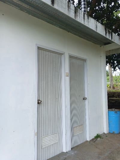 The image displays a segment of a white, single-story building located in Man Gyat, Myanmar. The building features two identical, light-colored vertically corrugated doors, each equipped with a silver-colored round doorknob and a rectangular louvered vent near the bottom. A light-colored electrical switch or outlet is positioned on the wall between the two doors. Above the doors, an overhang structure is visible, comprising a heavily stained white beam and a lighter-colored metal channel. The ground in the immediate foreground is a grey concrete slab, exhibiting minor cracks and supporting small patches of green weeds along the building's base on the right. A blue metal barrel with a black lid is situated on the concrete against the right side of the building. The background reveals green trees and foliage, with a distinct black and white checkered object (possibly a flag or cloth) partially visible amidst the upper tree canopy above the roofline. No individuals are present in the scene.