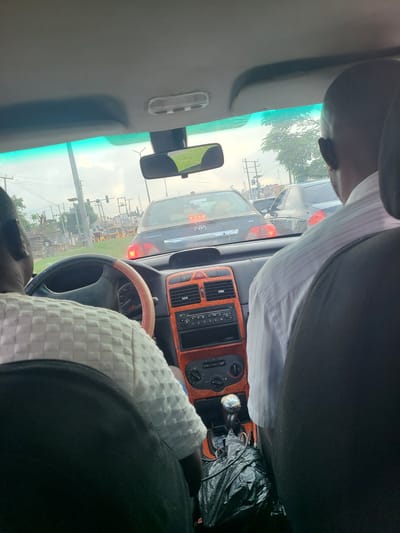 The image is a first-person perspective from inside a car in traffic in the city of Karu, Nigeria. The driver, an adult male with dark skin and a shaved head, is wearing a white patterned shirt and is visible from the back. To the right, another adult male with dark skin, wearing a light blue and white striped shirt, is also visible from the back. The car's dashboard features orange trim, a stereo system, air conditioning controls, and a gear shift. A black plastic bag is placed next to the gear shift. Outside the car, traffic is heavy, with several vehicles including a black Toyota Corolla and other cars and motorcycles visible. The sky is overcast, and there are trees and utility poles in the background. The overall impression is one of a typical urban traffic scene in Nigeria.