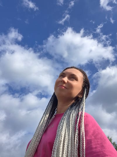 A woman with long, braided hair that is dark brown at the roots and transitions to white at the ends stands outdoors, looking upwards towards a blue sky with scattered white clouds. She is wearing a bright pink t-shirt. The image is shot from a low angle, with the sky occupying most of the frame. There are no discernible city landmarks or specific environmental features to indicate the location as Mozhary, Russia.