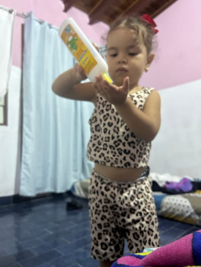 A toddler with a red bow in her hair is holding a bottle of lotion, posing for the picture. She is wearing a leopard print two-piece outfit. The room has pink walls, a white curtain, and blue tile flooring. The location is San Juan de Los Morros, Venezuela.
