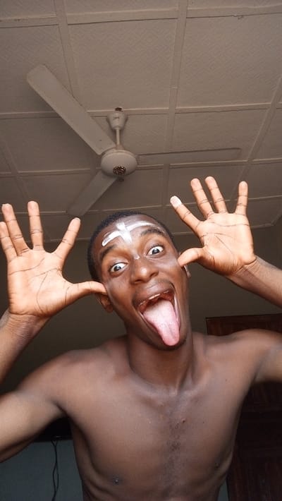 A shirtless dark-skinned man with a white cross painted on his forehead makes a silly face. He pulls his ears, widens his eyes, and sticks out his tongue. A ceiling fan is visible in the background, along with a textured ceiling and parts of a room.