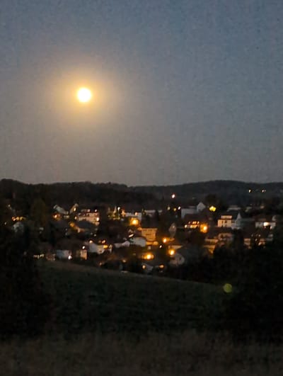 A hazy, blurred, full moon hangs in the twilight sky above a dimly lit residential area in St. Peter, Germany. The moon, emitting a soft yellow glow, is positioned in the upper left quadrant of the frame. Below the moon, a cluster of houses with illuminated windows forms a scattered pattern against a dark treeline. The foreground consists of a dark, out-of-focus grassy hill. The overall image is characterized by low light and a lack of sharp detail, suggesting it was taken with a camera in low-light conditions or with a wide aperture. No individuals or specific actions are discernible due to the image's blurriness and low resolution.