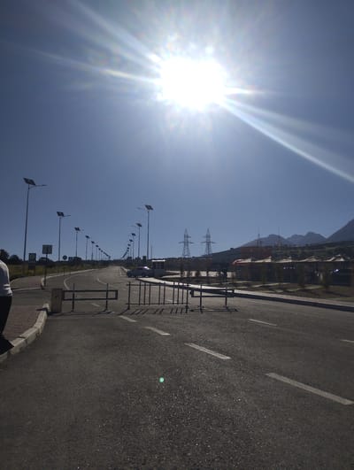 The image depicts a sun-drenched scene on a paved road, possibly in Dardar, Uzbekistan. A bright sun with strong lens flare dominates the upper portion of the sky, casting a harsh light. The road, marked with white lines, stretches into the distance, lined with numerous solar-powered streetlights. In the middle ground, a metal barrier is present on the road. Further back, several high-voltage power transmission towers are visible, along with some buildings and the silhouettes of mountains against the horizon. A white car is parked on the right side of the road. A person is partially visible on the left side of the frame, with their back to the camera. The environment appears to be semi-arid, with sparse vegetation visible along the roadside. The overall impression is one of an open, modern infrastructure development under a clear, bright sky.