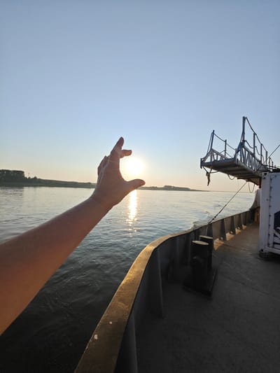 A person's arm and hand are extended in the foreground, fingers curved to appear to be holding the setting sun. The sun is low on the horizon, casting a bright reflection on the water. The scene is on a body of water, likely a river, with land visible in the distance. The perspective is from a boat, with the deck and railing of the vessel visible in the right side of the frame. A metal walkway extends from the boat into the air. The sky is clear and blue, transitioning to a lighter hue near the horizon. The location is the Angara River near Lesosibirsk, Russia.