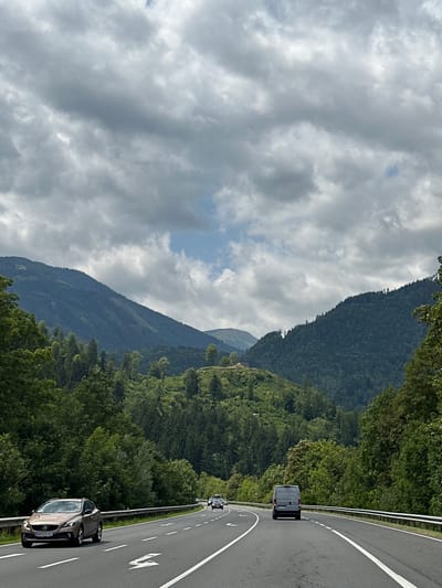 The image shows a highway in Pattendorf, Austria, on a cloudy day. The highway is paved and has multiple lanes with white lane markings. Several cars are visible on the road, including a beige Volvo SUV in the foreground on the left, and a silver van in the middle lane on the right. The highway is bordered by a metal guardrail and lush green trees. In the background, rolling hills covered with dense forests rise up to meet a dramatic, cloud-filled sky. The landscape suggests a mountainous or hilly region.